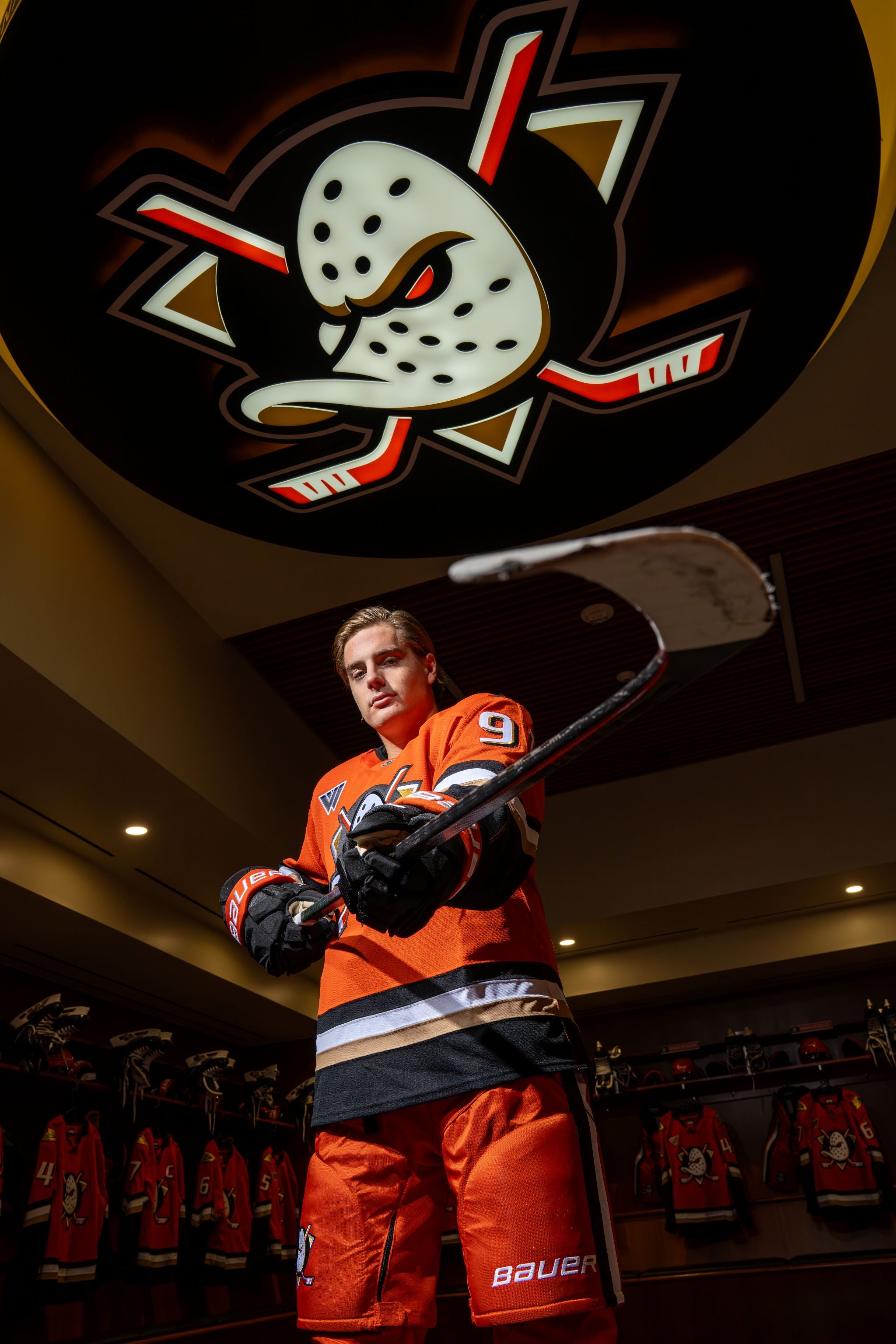 Leo Carlsson in the home uniform holding a hockey stick in the Anaheim Ducks' locker room at Honda Center.